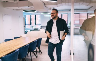 Gözlüklü, dizüstü bilgisayarlı, geniş bir ofiste yürüyen, modern iş merkezinde çalışan, başka yöne bakan ve gülümseyen pozitif bir erkek yönetici.