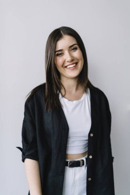Uzun saçlı, şık giyinmiş, gülümseyen ve kameraya bakan genç bir kadın stüdyoda beyaz arka planda duruyor.