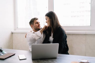 Yan görüntüde genç çift kucaklaşırken ve masada birbirlerine bakarken akıllı telefonlu dizüstü bilgisayarlarıyla evden ofise giderken güneş ışığı penceresini aydınlatıyorlardı.