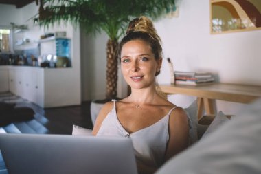 Keyifli, genç, uzaktan kumandalı bir kadın. Gündelik kıyafetlerle kameraya bakıyor. Dizüstü bilgisayar projesinde çalışıyor ve evde çalışırken gülümsüyor.