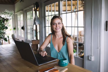 Keyifli, uzaktan kumandalı, günlük kıyafetli, gülümseyen ve kameraya bakan genç bayan bir yandan da dizüstü bilgisayarda bir fincan kahveyle çalışıyor.