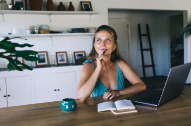 Düşünceli, gündelik giysiler içinde genç bir bayan. Masada açık bir defterle oturuyor ve elinde kalemle başka yere bakıyor.