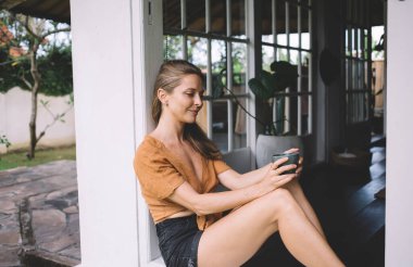 Rahat giyinen, gülümseyen ve terasta oturan, hafta sonu dinlenirken bir bardak lezzetli içecekle yetinen genç kadın.