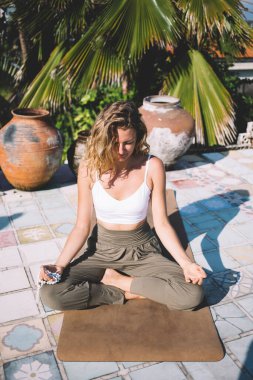 Gün ışığında gözleri kapalı genç bir kadın elinde dua boncuklarıyla mindere oturmuş palmiye ağaçlarının yakınındaki tropik tatil köylerinde meditasyon yaparken parmakları Mudra 'nın içinde.