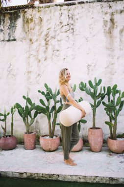 Çıplak ayaklı, sıska bir kadın. Kaktüs bitkilerinin yanında duruyor. Yoga çantasıyla eski püskü duvarlara bakıyor.