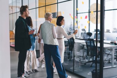 Çok ırklı iş adamları fikirlerini paylaşıyor ve cam duvara notlar yazarken aynı zamanda yeni girişimlerin iş planlarını analiz ediyor ve modern ofiste işbirliği yapıyorlar.