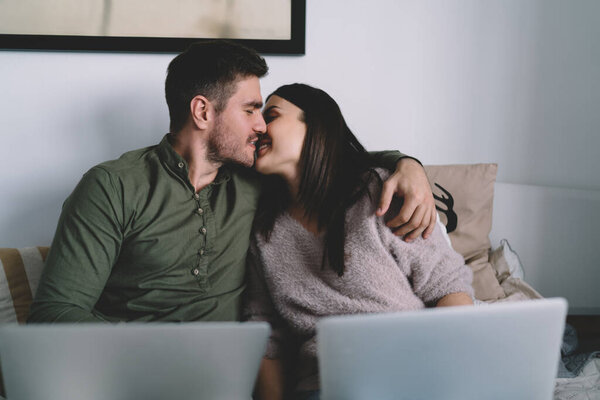 Young cute hetrosexual smiling couple having romantic moment at home while kissing and cuddling sitting on bed together with laptops