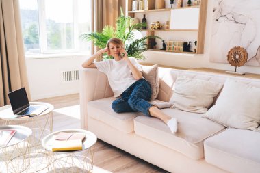 Kot pantolonlu ve tişörtlü yetişkin bir kadın kanepede dinleniyor ve cep telefonuyla konuşurken elini uzatıyor ve hafta sonu evdeki oturma odasına bakıyor.