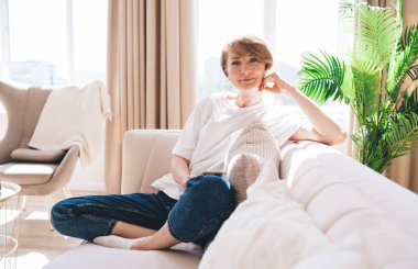 Uzun boylu, kısa saçlı, kanepede dinlenen, bacak bacak bacak üstüne atmış ve hafta sonu evde dinlenirken eline yaslanan kaygısız bir kadın.