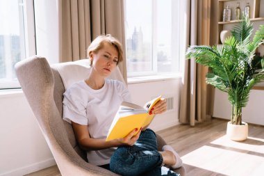 Odaklanmış sarışın kadın rahat koltuğa oturuyor ve evde boş vakti varken dikkatlice kitap okuyor.