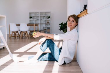 Orta yaşlı kadın, gündelik giysiler içinde yerde oturuyor ve elleri dizlerinin üstünde, beyaz duvarda elmalarıyla dinleniyor.