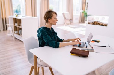 Klavyeye bilgi yazarken dizüstü bilgisayarlı ve akıllı telefonlu, günlük giysiler içinde kadın serbest çalışanın yan görüntüsü