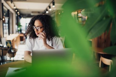 Bulanık yeşil yaprakların arasından gözlüklü genç kız öğrencinin cep telefonuyla konuşurken yüzünü çevirmesi ve kafede çalışırken dizüstü bilgisayarlı kalem panosuyla oturması.