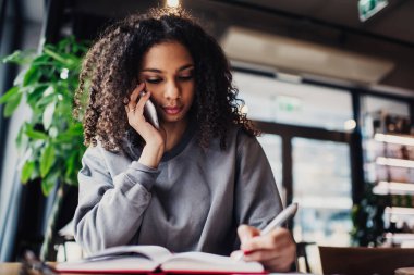 Kafedeki cep telefonu konuşmaları sırasında not defterine kalemle not alarak masada oturan konsantre olmuş Afro-Amerikan kız öğrencinin düşük açısı.