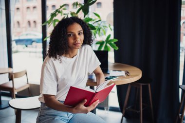 Afrika kökenli Amerikalı bir kız öğrenci elinde dizüstü bilgisayar gözlüklü akıllı telefon panosuyla masada oturuyor ve gün içinde kafede ödev detaylarını incelerken kameraya bakıyor..