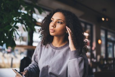 Düşük açılı, genç, düşünceli, etnik bir kadın. Cep telefonu elinde, başka bir yere bakıyor. El ele oturuyor, şakak kıllarına dokunuyor.