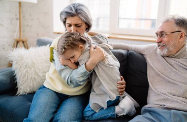 Mutlu son sınıf çifti küçük kızla koltukta otururken yaşlı kadın üzgün torununu kucaklıyor ve evde birlikte vakit geçirirken kafasından nazikçe öpüyor.