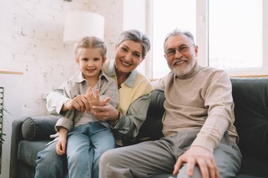 Mutlu büyükanne ve büyükbabanın aile portresi. Torunuyla kucaklaşıyorlar. Kanepede oturup kameraya bakarken gülümsüyorlar.
