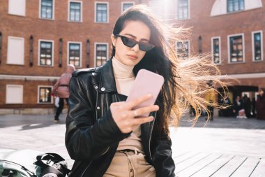 Deri ceketli, güneş gözlüklü, cep telefonuyla internette sörf yapan, haftasonu bankın yanında dinlenen şık bir bayan.