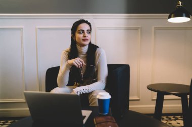 Kendine güveni olan genç bir kadın elinde gözlük tutarken ve masadaki koltukta dizüstü bilgisayarı ve kahveyle otururken yan lambanın yanındaki oturma odasında dinleniyor.