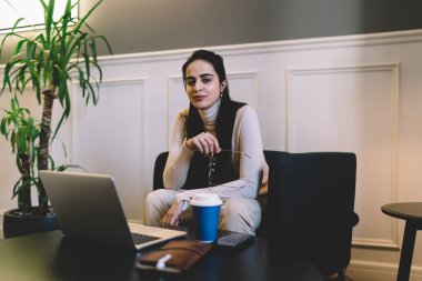 Elinde gözlüklerle masada dizüstü bilgisayarlı kahve fincanı ile oturan ve oturma odasındaki saksı bitkisinin yanında dinlenirken kameraya bakan pozitif genç bir bayan serbest yazar.
