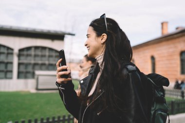 Deri ceketli, şehir caddesinde durup cep telefonuyla selfie çeken genç bir kadının yan görüntüsü.