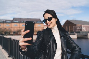 Deri ceketli ve güneş gözlüklü genç bir kadın nehir kenarında dururken cep telefonuyla kendi portresini çekiyor.