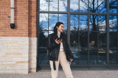 Siyah ceketli neşeli genç bir bayan elinde cep telefonuyla restoranda oturan ve mavi gökyüzü ışıklarını ve bulutlarını gösteren yansıtıcı gözlükleriyle müşterilere doğru bakıyor.
