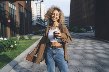 Elinde kahve fincanı ve not defteriyle kaldırımda dikilirken gülümseyen ve kameraya bakan Afro-Amerikan kız öğrenci.