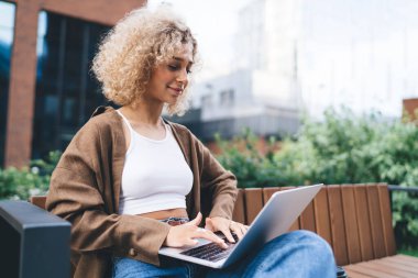 Çevrimiçi dizüstü bilgisayarla proje üzerinde çalışırken, tahta bankta oturan, gündelik kıyafetleri ve kıvırcık saçları olan, pozitif, genç, etnik, serbest bir kadın.