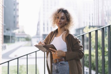 Kıvırcık saçlı, gülümseyen ve elinde kalemle çitin yanında durup kameraya bakan Afro-Amerikan genç bir kız öğrenci.