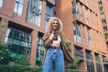 Sıradan kıyafetlerle gülümseyen siyah bir kadının kulaklıkla müzik dinlemesi ve şehir caddesinde akıllı telefon kullanırken kameraya bakması.