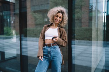 Pozitif Afro-Amerikan kadın kıvırcık saçlı, gülümseyen ve kameraya bakan şehir sokaklarındaki modern binanın cam duvarına yaslanmış.