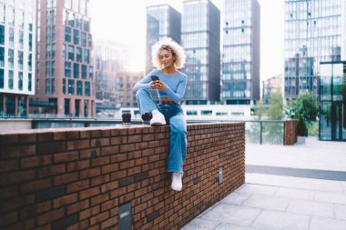 Genç Afro-Amerikan kadın bacağı duvarda oturuyor ve dizlerine yaslanıp telefon ekranına bakarken cep telefonunu karıştırıyor.