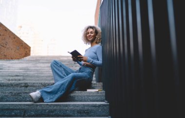 Kameraya bakıp metal çitlere yaslanırken elinde kitapla basamaklarda oturan neşeli, Afro-Amerikan genç kız öğrencinin yan görüntüsü