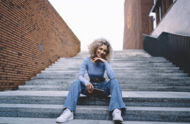 Pozitif, neşeli, genç, Afro-Amerikalı bir kadın. Basamaklarda oturuyor, başı öne eğik ve kameraya bakıyor.