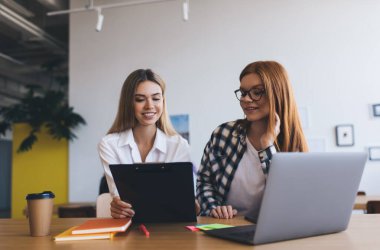 Gündelik kıyafetleri olan pozitif kadın çalışanlar, modern minimalist tarz ofis içinde netbook ve kırtasiye malzemeleriyle masa başında otururken iş projeleriyle ilgili kağıtlarla birlikte çalışıyorlar.