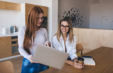Ellerinde dizüstü bilgisayarı olan ve birlikte ekrana bakan genç ve pozitif bayan çalışanlar. Bu sırada gözlemi işaret ediyorlar ve ofiste çalışırken masada dizüstü bilgisayarlı akıllı telefon ve bir fincan kahveyle duruyorlar.