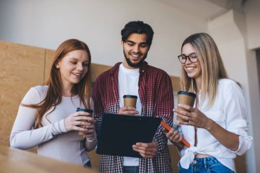 Ellerinde kahve fincanlarıyla ayakta duran ve modern ofisteki projenin eğlenceli bilgilerini bulurken erkek meslektaşlarının panolarına bakan bir grup pozitif iş arkadaşı.