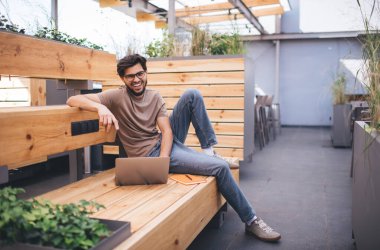 Sırıtkan sakallı, tahta bankta dizüstü bilgisayarlı kalemle teras bahçesindeki kafede dinlenen ve gündüz vakti yeşil bitkilerin yanında vakit geçirirken kameraya bakan bir vücut.