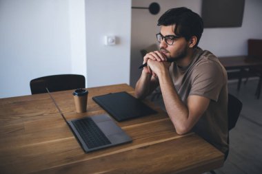 Düşünceli, gözlüklü, elleri çenesine değen, dirsekleriyle masaya yaslanan ve modern çalışma alanındaki dizüstü bilgisayara bakarken bir fincan kahve grafikleri tabletine bakan serbest bir erkeğin yan görüntüsü.
