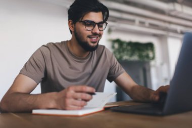 Günlük giysiler ve gözlüklerle serbest çalışan genç, pozitif sakallı bir erkek. Gündüz vakti yalnız oturuyor ve netbook 'ta geziniyor.