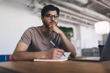 Gözlüklü, elleri çene hizasında, dizüstü bilgisayarlı kahve fincanı ile masaya otururken kameraya bakan ve bulanık beyaz binadaki modern çalışma alanında not defterine notlar yazan kendine güvenen genç bir serbest erkek.
