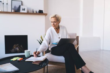 Yoğunlaşmış kadın girişimci masada dizüstü bilgisayarla bacak bacak üstüne atarak oturuyor ve ekranda bilgi okurken elinde kalemle deftere bakıyor.