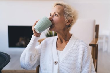 İyimser yaşlı kadın, evde şömineyle oturma odasında bardaktan sıcak içecek içerken yukarı bakıyor.