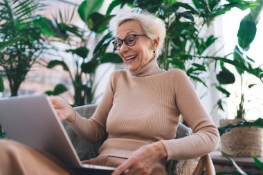 Boğazlı kazaklı, kısa sarı saçlı, gülen yaşlı kadın. Ekoseli hasır sandalyede oturuyor ve yeşil bitkilerin arasında dizüstü bilgisayarla komik filmler izliyor.