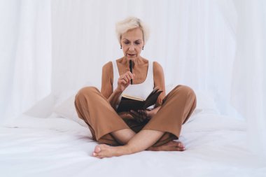 Uzun boylu, çıplak ayaklı, gri saçlı, bacak bacak üstüne atmış, çenesine kalemle dokunmuş ve hafta sonu yatak odasında günlük okuyan olgun bir kadın.