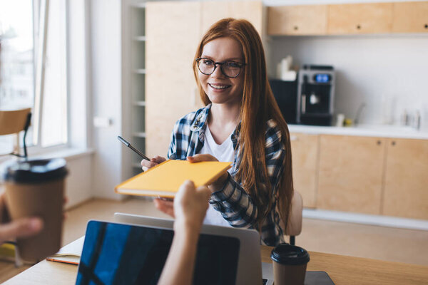 Позитивная молодая женщина в очках сидит с неузнаваемым коллегой за столом с кофейной чашкой для ноутбуков и передает книгу, глядя друг на друга в офисной столовой во время кофе-брейка
