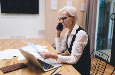 Resmi elbiseli, meşgul, yaşlı bir iş kadınının modern ofiste el ele dokümanları incelerken dizüstü bilgisayarla masaya oturması ve cep telefonuyla konuşması.