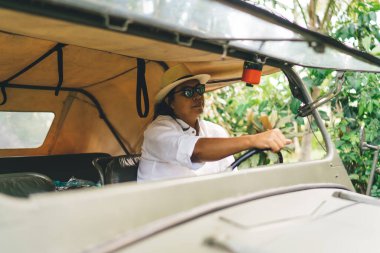 Asyalı erkek gezginin günlük kıyafetleri ve şapkasıyla güneş gözlüklü bir minibüsün sürücü koltuğunda oturması ve yolculuk sırasında gözlerini kaçırması.